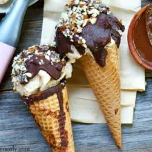 Two vegan drumsticks ice cream cones sit on a wooden counter next to chocolate syrup and a pink ice cream scoop.