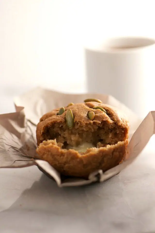 A muffin is torn in half and a pat of melted butter is inside. It sits in a tall brown paper. A mug of tea is behind it.