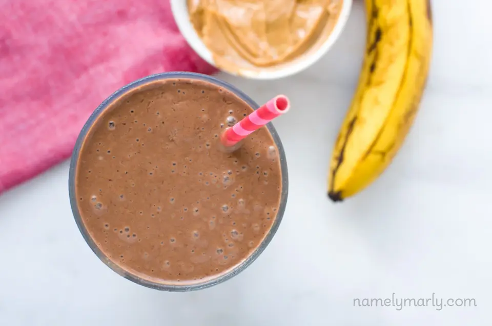 A top-down shot looking at chocolate peanut butter protein in a glass. There's a pink straw in the smoothie. There's a bowl of peanut butter and a ripe banana sitting next to it.