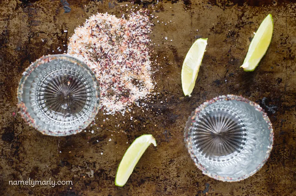 Looking down on two glasses with edges dipped in a spicy, salty mixture sitting next to lime wedges.