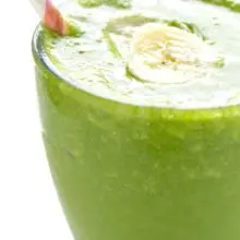 A a clear glass is full to the rim with green banana smoothie. A pink and white paper straw is on the side, and a slice of banana is in the middle of the smoothie.