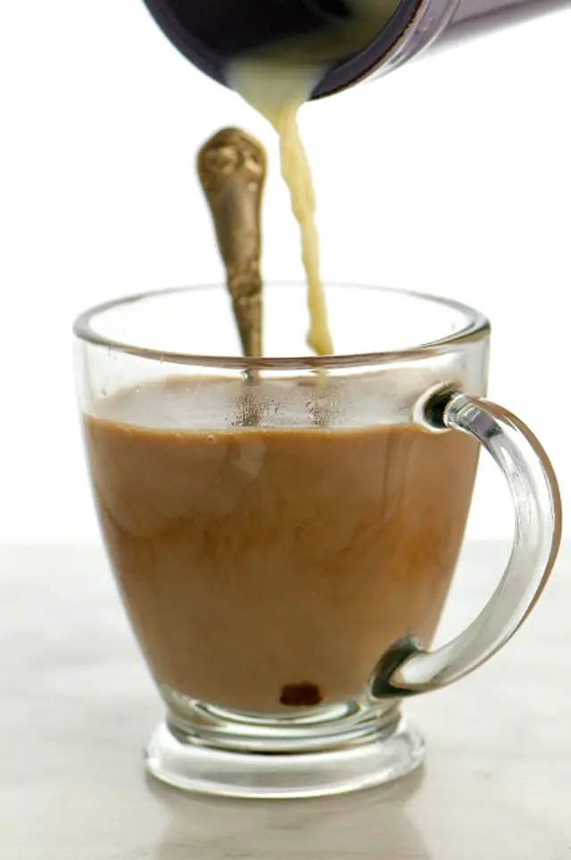 A container pours plant-based milk into a cup of cocoa with a spoon in it.