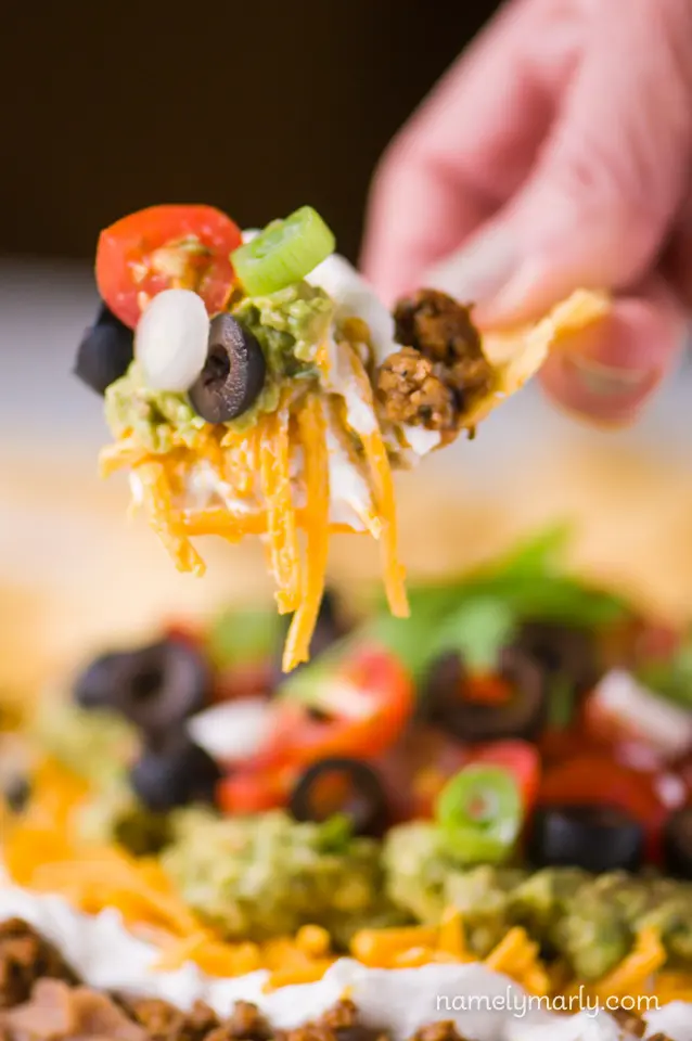 A hand holds a tortilla chip loaded with vegan seven layer dip. The plate of dip is sitting in the background.