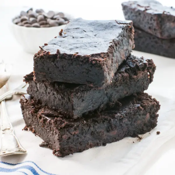 Three slices of black bean brownies are stacked on top of each other with a bowl of chocolate chips behind them.