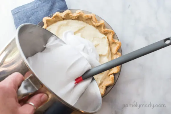 A hand holds a mixing bowl and is pouring meringue over the top of a pie.