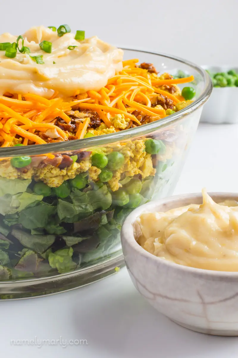 A layered salad is topped with vegan ranch dressing with a bowl of frozen peas behind it.
