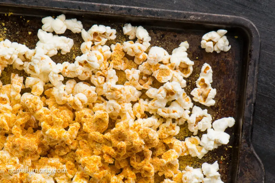 A close-up shot of popcorn on a baking sheet with lots of cheesy nutritional yeast flakes sprinkled on top.