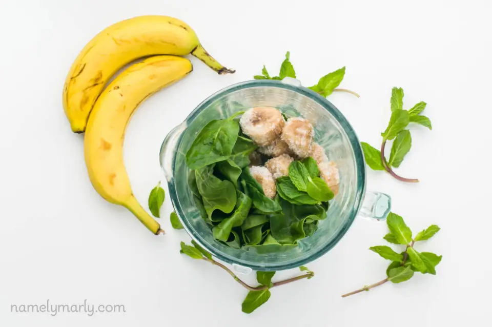 Spinach and frozen bananas in a blender with mint sprigs and a couple of bananas on the counter behind it.