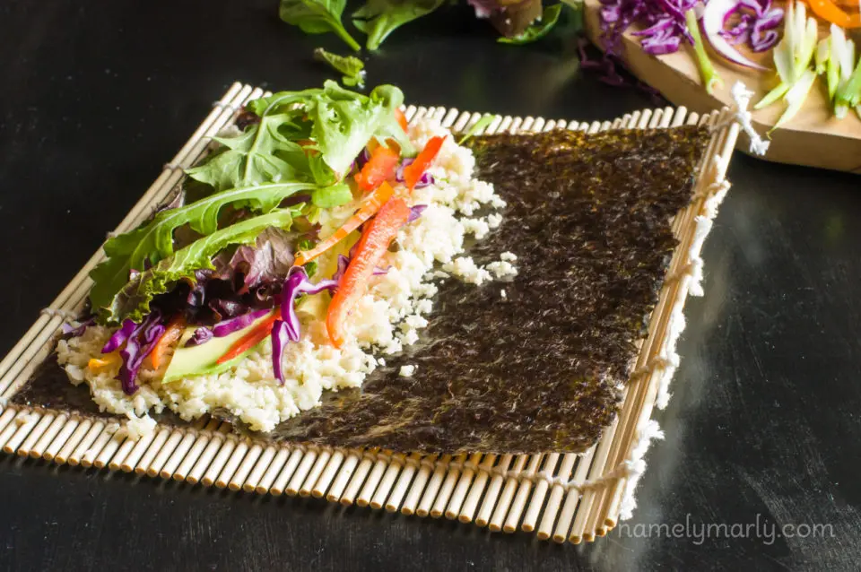 A sheet of nori is on a bamboo mat, with cauliflower rice topped with sliced veggies.