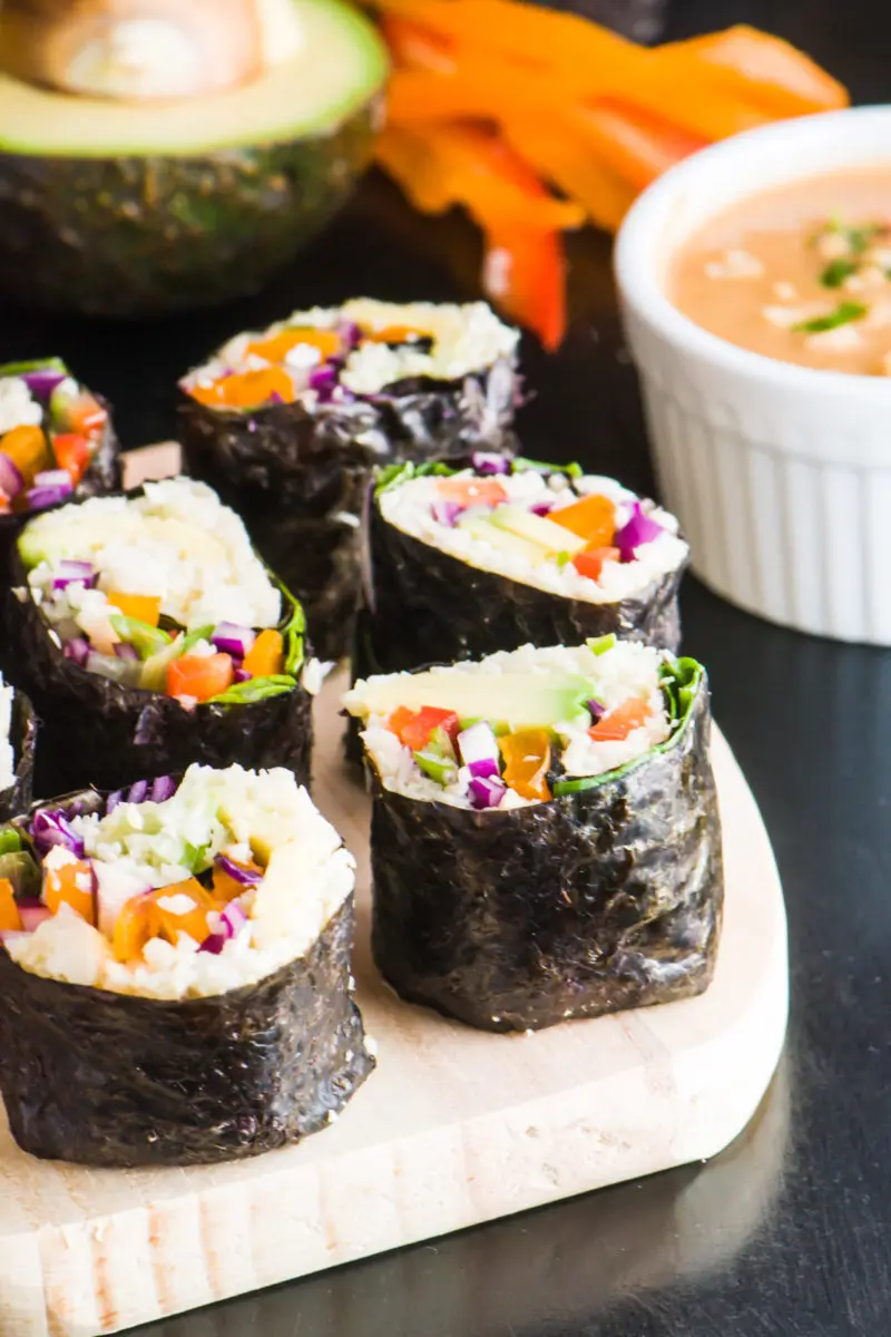 Several slices of vegan sushi with the dark nori on the outside sits by a bowl of spicy sweet peanut butter sauce.