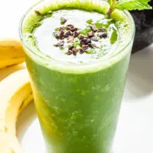 A green smoothie in a tall glass has cacao nibs and mint sprigs in it. There are bananas next to it and an avocado behind it.