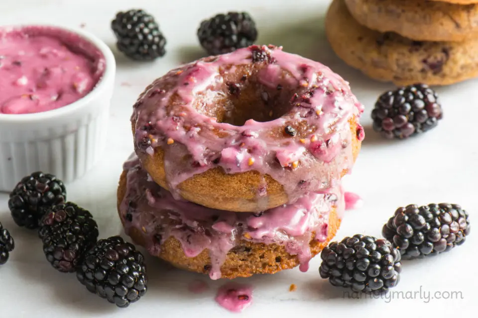 Two glazed blackberry donuts surrounded by fresh blackberries and a bowl of blackberry glaze.