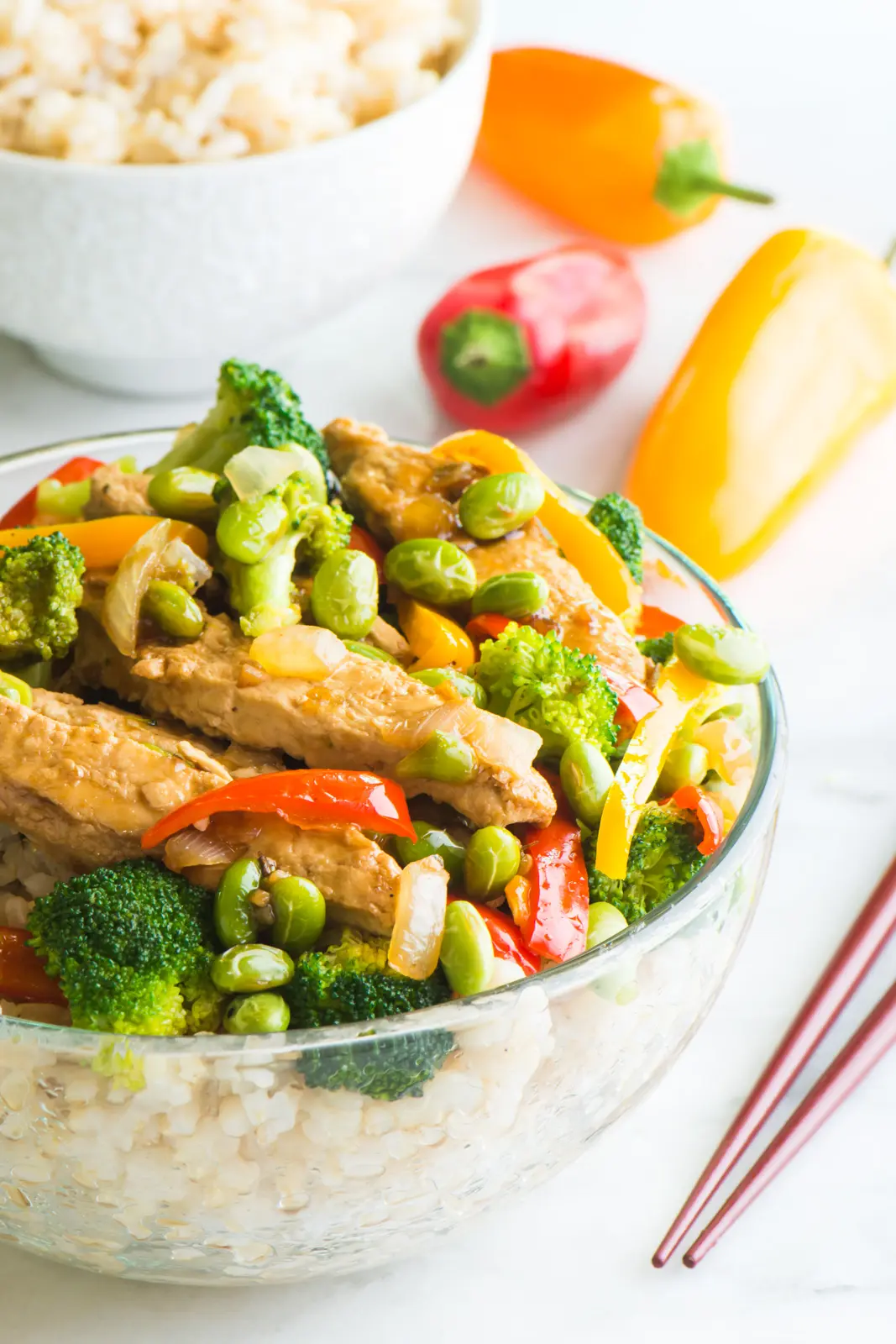 A vegan teriyaki chicken and edamame rice bowl sits next to chopsticks with a bowl of rice behind it and mini bell peppers. 