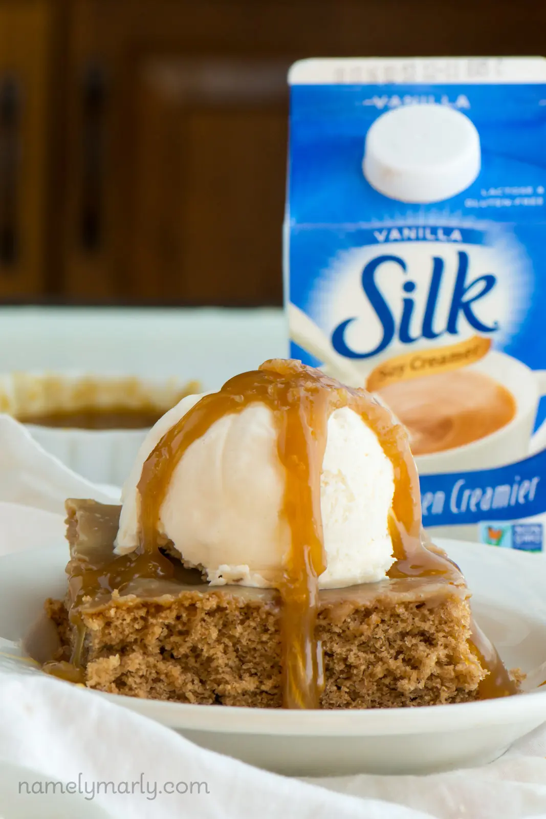 A slice of vegan cinnamon roll poke cake with ice cream and caramel sauce sits in front of a bowl of caramel sauce and a carton of soy milk.
