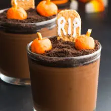 Two small glasses hold chocolate pudding with candy pumpkins and a RIP graham cracker tombstone. These are vegan Halloween Pudding cups to serve at parties.