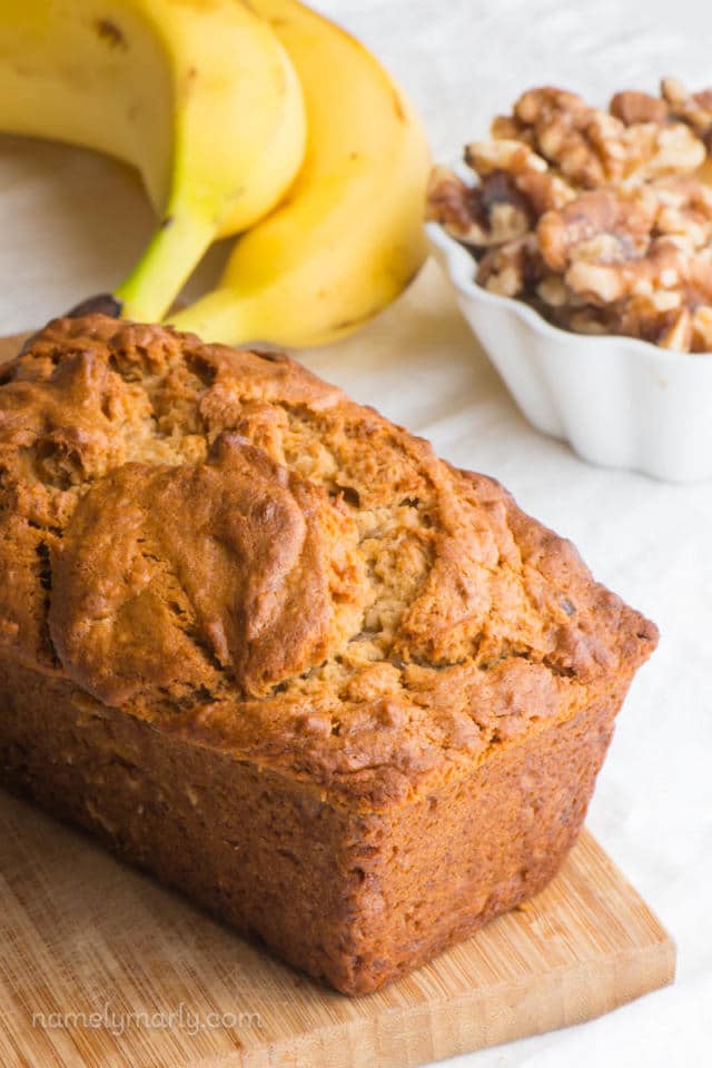 A loaf of banana bread sits on a cutting board with a bowl of walnuts and a banana behind it.