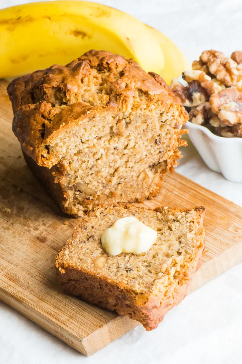 A slice of vegan banana bread with a pat of butter on it sits in front of the rest of the loaf.