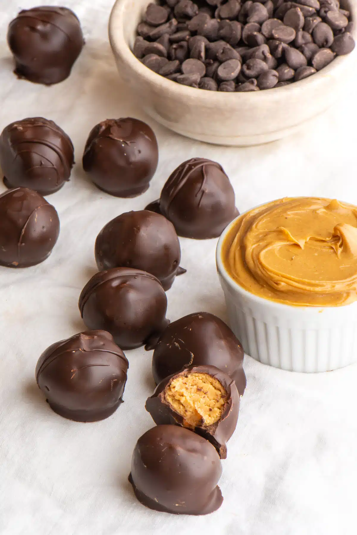 Several peanut butter balls, one with a bite taken out, sit next to a bowl of peanut butter and another bowl of chocolate chips.