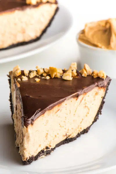 A slice of peanut butter pie sits on a plate. there's a bowl of peanut butter behind it along with another slice of pie on a plate.