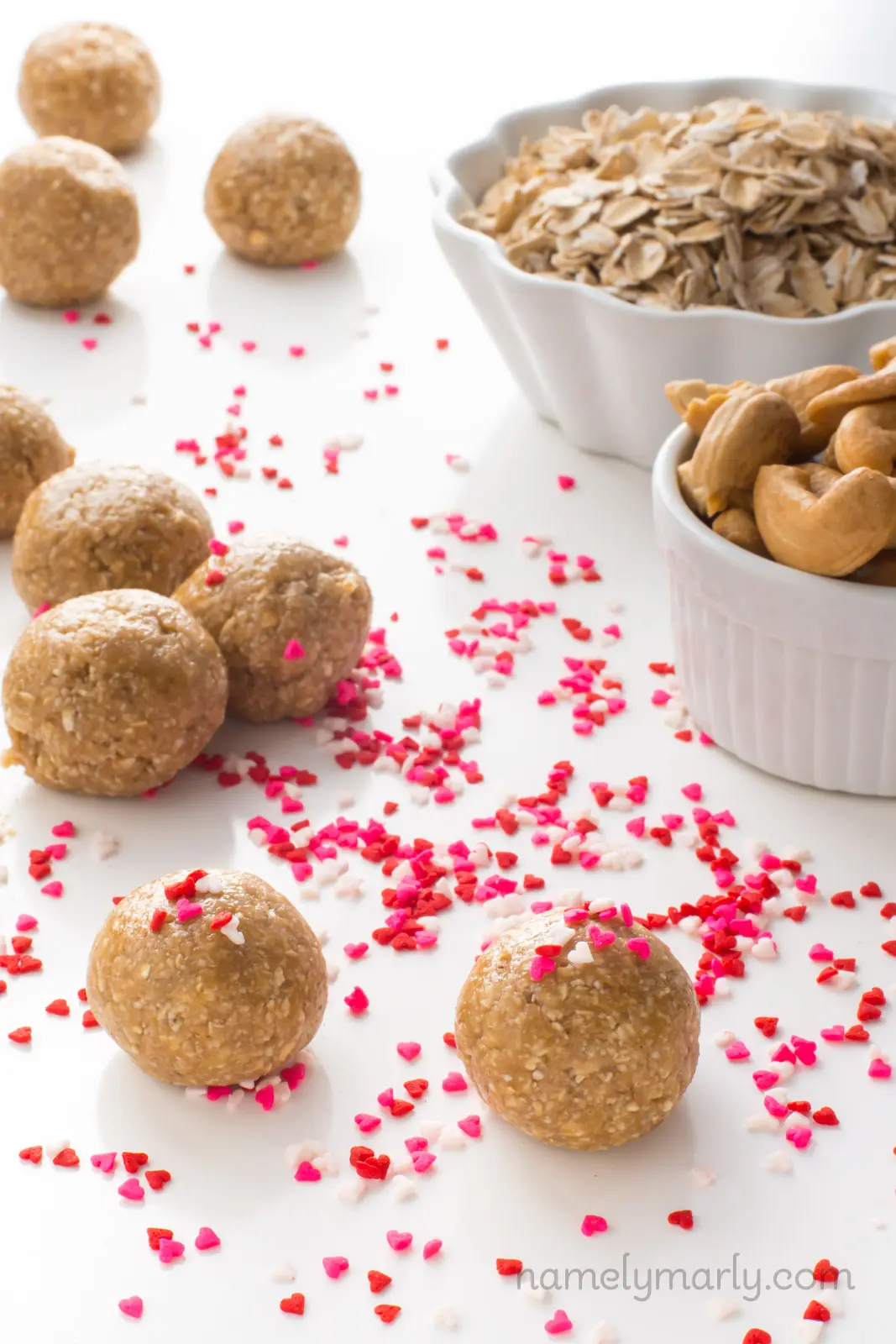 Several sugar cookie energy bites sit on a table. There are pink, red, and white heart-shaped sprinkles all around the table and on some of the energy bites. Beside them are two bowls, one with cashews and one with oatmeal.