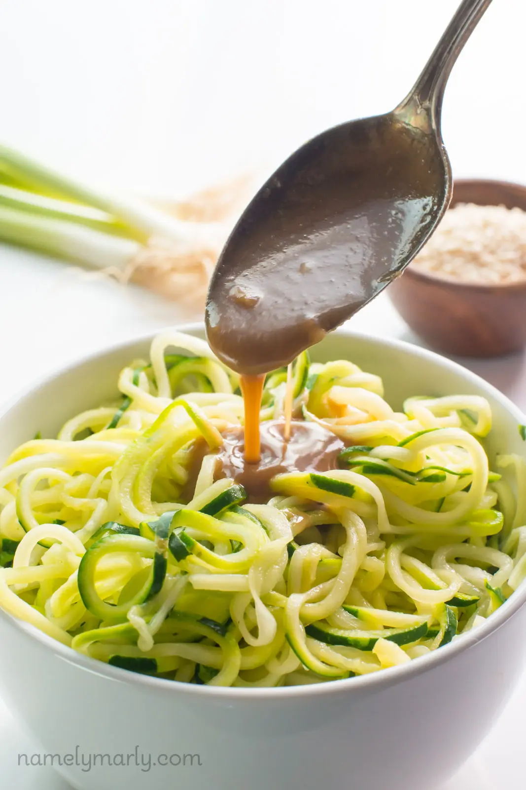 A spoon drizzles sauce over spicy sesame zoodles with green onions and sesame seeds behind it.