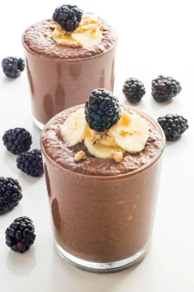 A serving glass holds vegan chocolate chia pudding topped with sliced bananas and fresh blackberries. Another glass and more blackberries is behind it.
