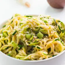 A bowl of spicy sesame zoodles with sesame seeds sits in front of green onions and a bowl of more sesame seeds.