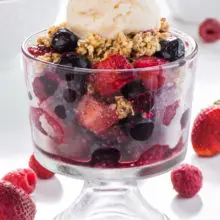 A bowl of vegan no-bake cherry fruit crisp in a serving dish with ice cream on top and other toppings behind it.