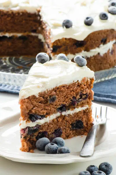 A slice of layered vegan Blueberry Cake on a plate in front of the rest of the cake.
