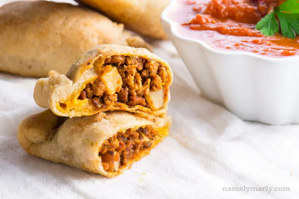 An empanada is cut and half and stacked. It sits next to red sauce and more empanadas behind it.