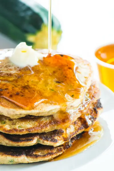 A stack of zucchini pancakes has maple syrup being drizzled over the top. More maple syrup and a zucchini is in the background.