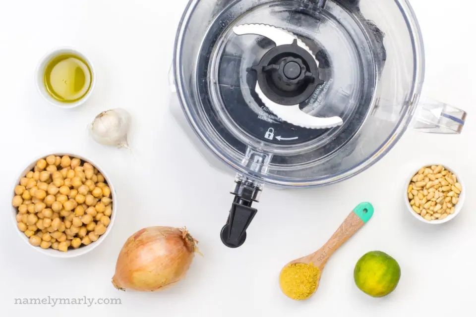 Looking down on a food processor with ingredients around it such as bowls of olive oil, chickpeas, a lime, an onion, garlic, and more.