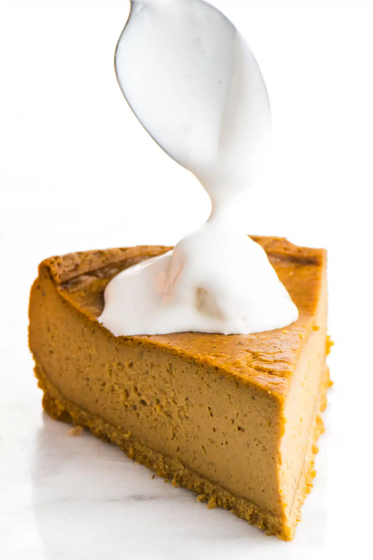 A spoon hovers over a slice of cheesecake, drizzling whipped cream over the top.