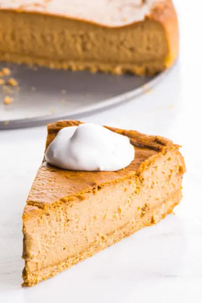 A slice of Vegan Pumpkin Cheesecake is served with vegan whipped cream on top. The rest of the cheesecake is behind it.