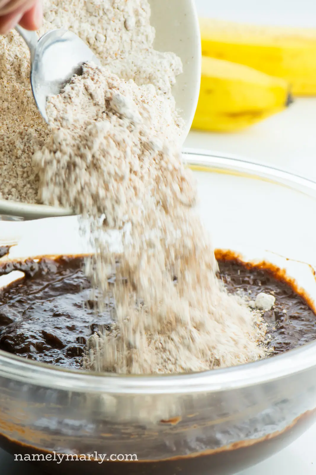 Whole wheat pastry flour is being poured into a a bowl of chocolate batter. Bananas are in the background.