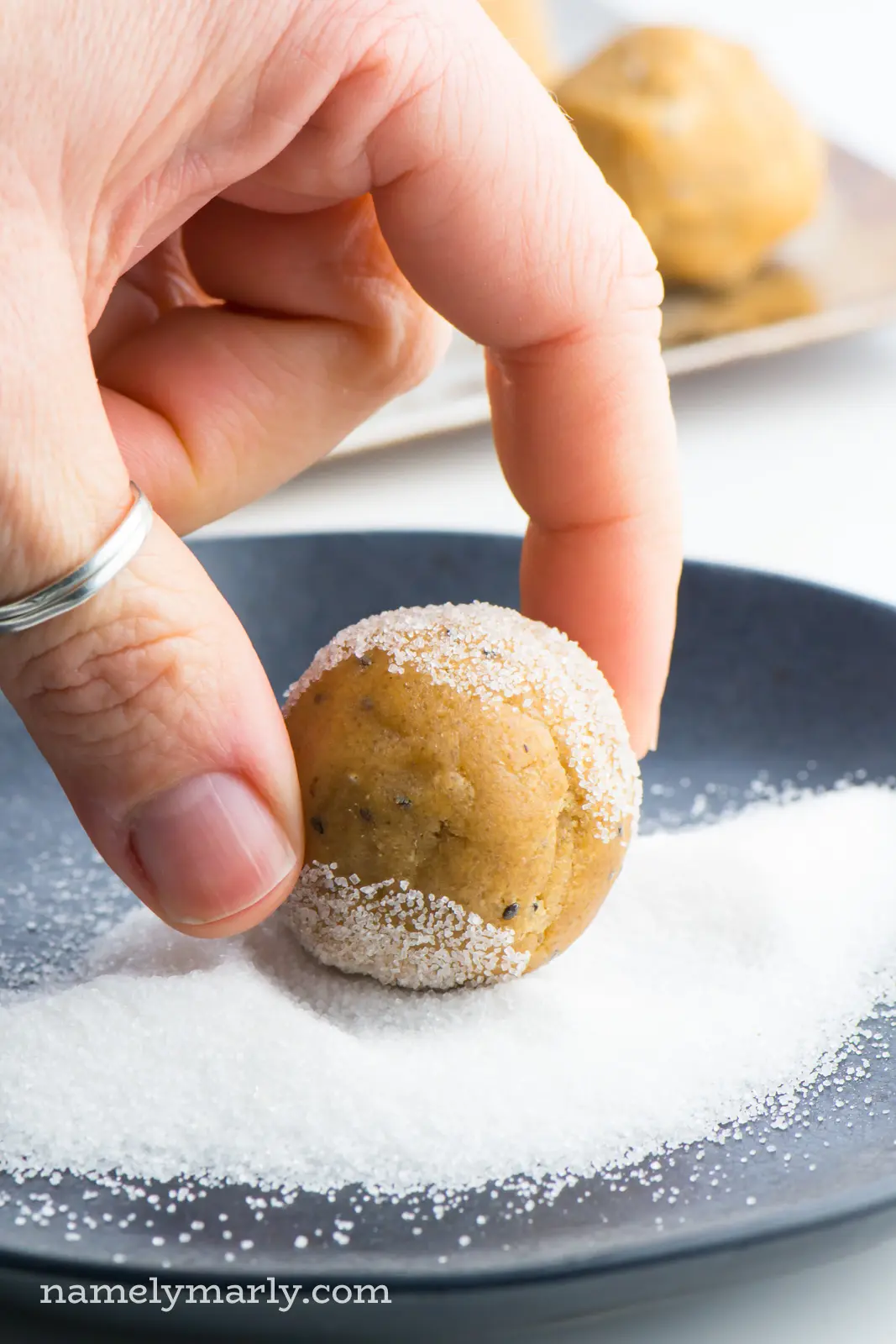 A hand is holding a vegan peanut butter cookie dough ball over a plate of sugar. The cookie dough ball has been partially rolled in the sugar.
