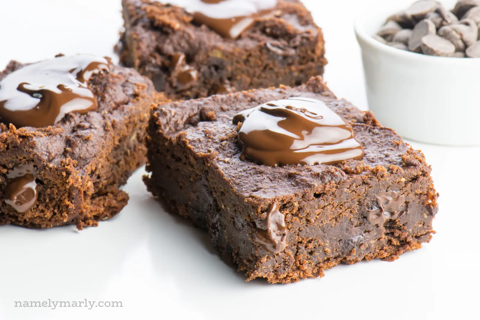 Three slices of chocolate banana brownies sit next to each other, each with a dollop of melted chocolate over the top. A white bowl full of chocolate chips sits nearby.