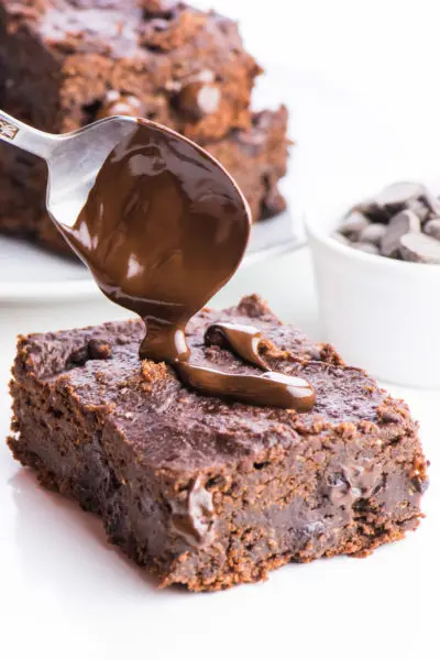 A spoon hovers over these chocolate banana brownies, drizzling melty chocolate over the top. A bowl of chocolate chips sits on the right and more slices of brownies are in the background.