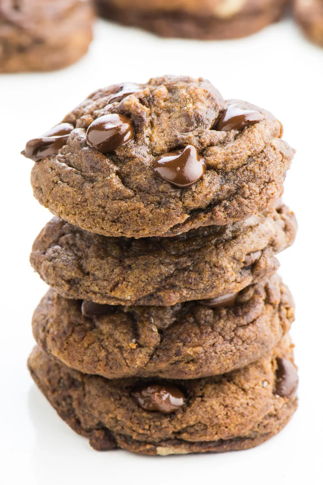 A stack of four vegan chocolate chocolate chip cookies sit one on top of the other. The chips are still warm and shining in the light. There are more cookies in the background.