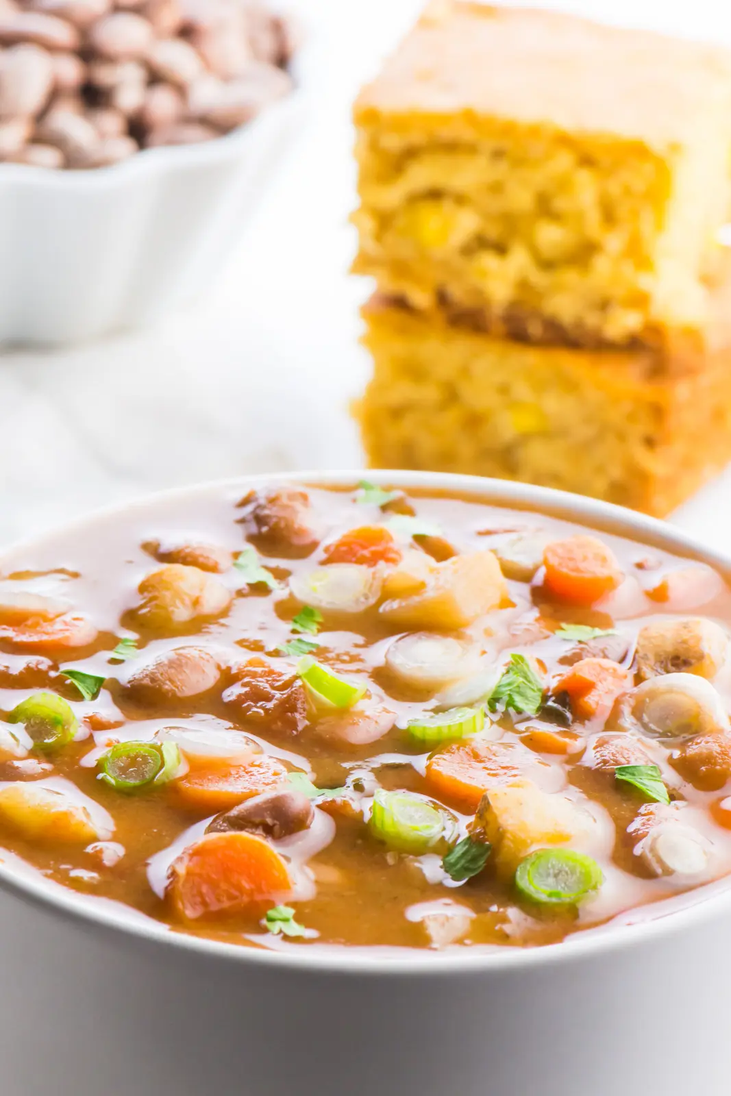 A bowl of pinto bean soup is front in center in this photo. The light is shining on all the ingredients in the bowl, including the slices of carrots, potatoes, and beans. There is a stack of cornbread behind the soup, and a bowl of dried pinto beans behind that.