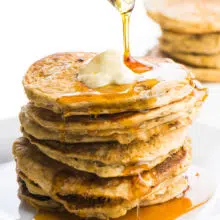 A stack of pancakes without eggs is sitting on a plate. The top one has a pat of melty vegan butter. A glass pitcher is pouring maple syrup and it is dropping down the sides of the pancakes. There are more pancakes in the background.