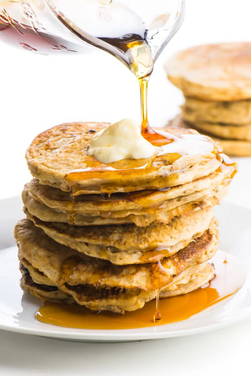 A stack of pancakes without eggs is sitting on a plate. The top one has a pat of melty vegan butter. A glass pitcher is pouring maple syrup and it is dropping down the sides of the pancakes. There are more pancakes in the background.