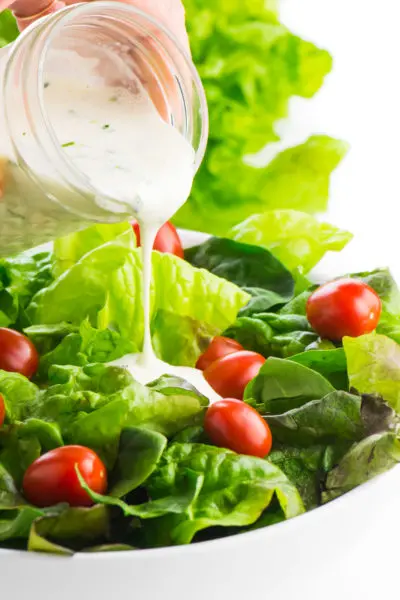 A mason jar full of vegan ranch dressing is pouring the dressing over a leafy, green salad with lots of bright red cherry tomatoes.