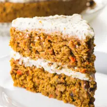 A slice of healthy carrot cake sits on a plate in front of the whole cake behind it.