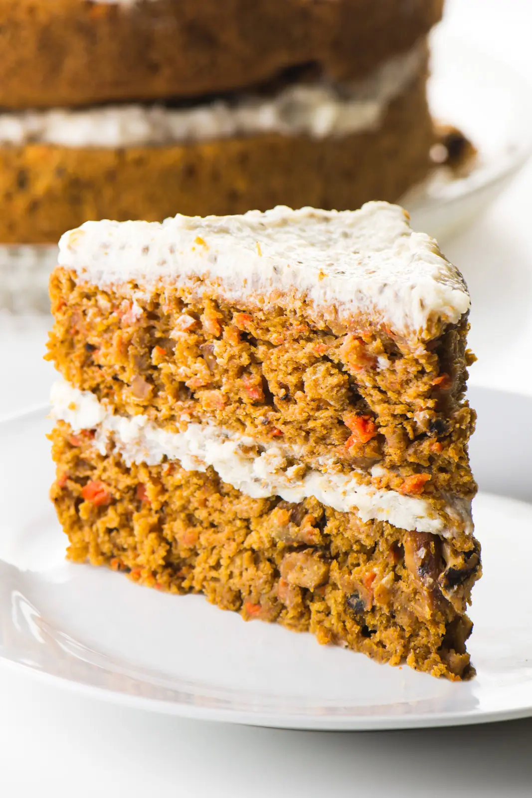 A slice of healthy carrot cake sits on a plate in front of the whole cake behind it.