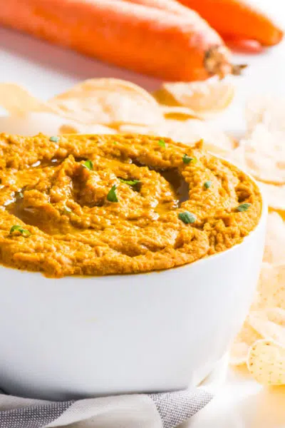 A bowl of carrot hummus is surrounded by chips with carrots in the background.