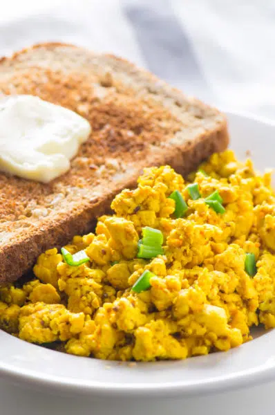 A piece of toast with a pat of melty vegan margarine sits behind a serving of tofu scramble.