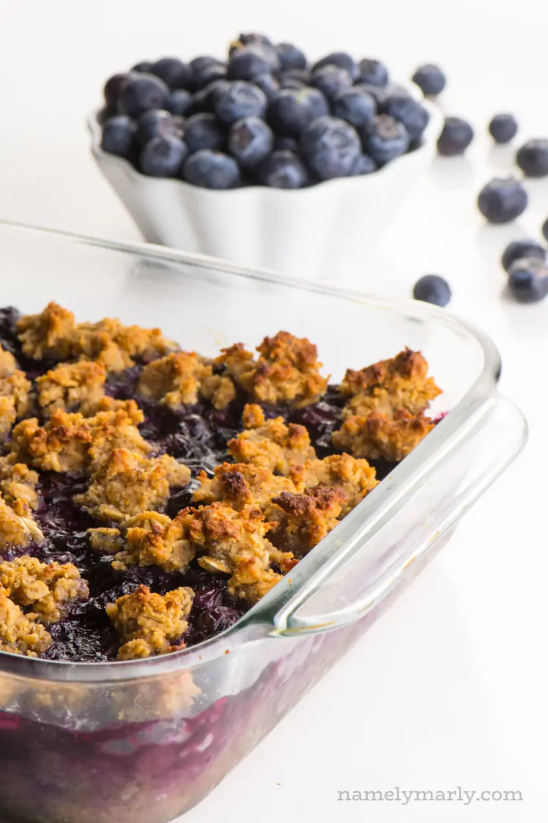 A baking dish full of blueberry crumble bars sits next to a bowl of fresh blueberries.