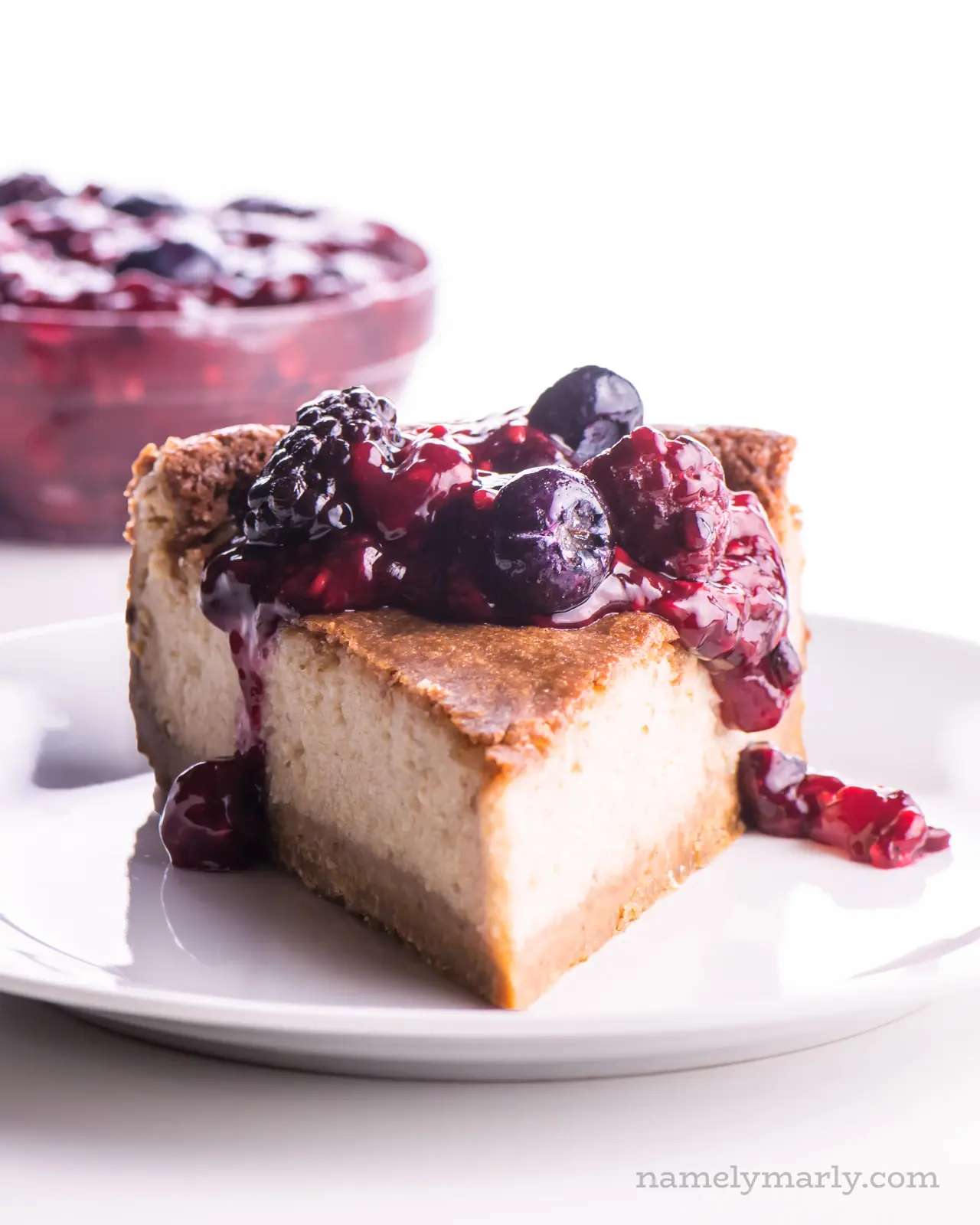 A slice of cheesecake with berry drizzle over the top and more berry topping in a bowl behind it.
