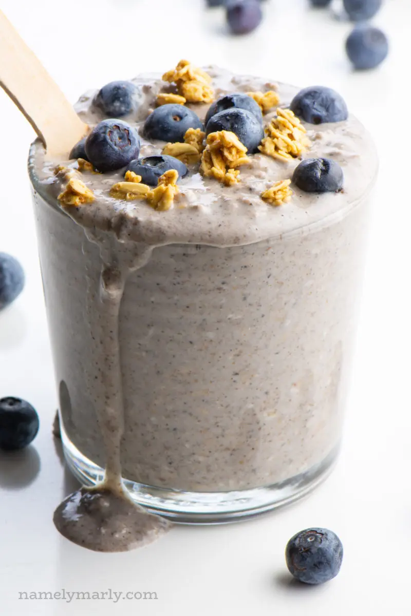 A single glass of overnight oats is topped with fresh blueberries and granola and has a spoon in it.